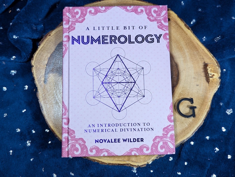 May include: A book titled "A Little Bit of Numerology" with a geometric design on the cover. The book is on a wooden slice with a dark blue background. The author's name is Novalee Wilder.