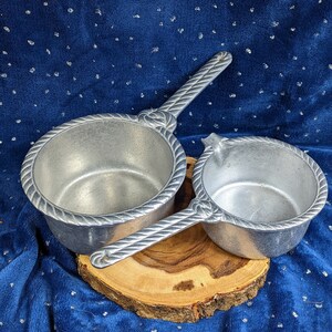 May include: Two silver aluminum serving dishes with rope-like handles. The dishes are on a wooden base and have a blue background.