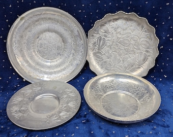 May include: Four silver aluminum serving trays with embossed designs. The trays are round and have a raised rim. The designs include a floral pattern, a geometric pattern, and a leaf pattern.