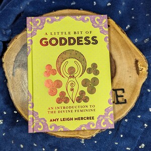 May include: A book titled "A Little Bit of Goddess" with a yellow cover and pink and gold accents. The book rests on a wooden slice against a blue background. The book's cover features an illustration of a female figure.
