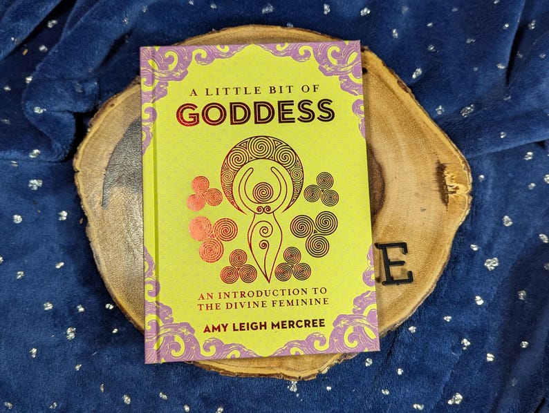 May include: A book titled "A Little Bit of Goddess" with a yellow cover and pink and gold accents. The book rests on a wooden slice against a blue background. The book's cover features an illustration of a female figure.