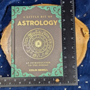 May include: A hardcover book titled "A Little Bit of Astrology" with gold lettering and a zodiac wheel illustration. The book is approximately 7 inches tall and 5 inches wide, with a dark gray cover and green decorative borders.