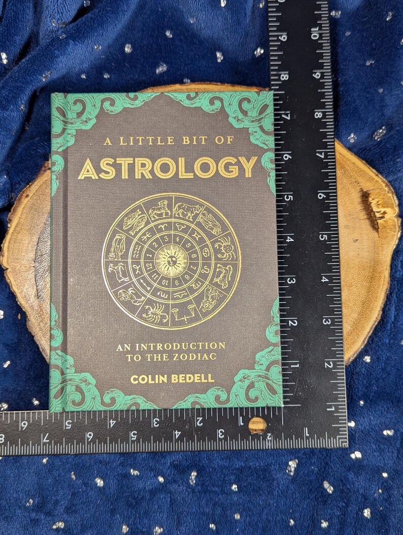 May include: A hardcover book titled "A Little Bit of Astrology" with gold lettering and a zodiac wheel illustration. The book is approximately 7 inches tall and 5 inches wide, with a dark gray cover and green decorative borders.