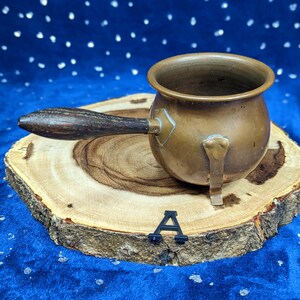 May include: A small, antique copper pot with a wooden handle. The pot is round with a spout and a small metal foot. It sits on a wooden slice against a blue background.