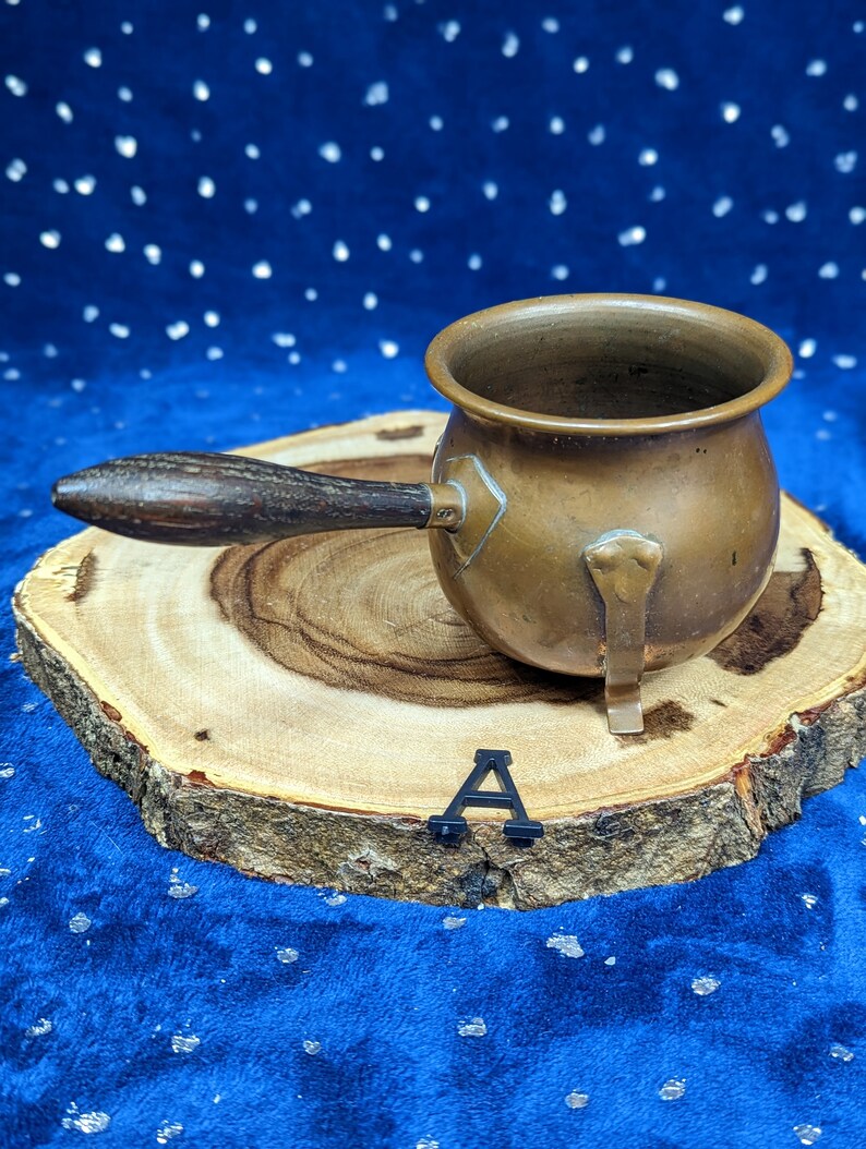 May include: A small, antique copper pot with a wooden handle. The pot is round with a spout and a small metal foot. It sits on a wooden slice against a blue background.