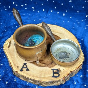 May include: Two small copper pots with wooden handles rest on a wooden slice. One pot is filled with blue and green material. Black letters A and B are placed near the pots.