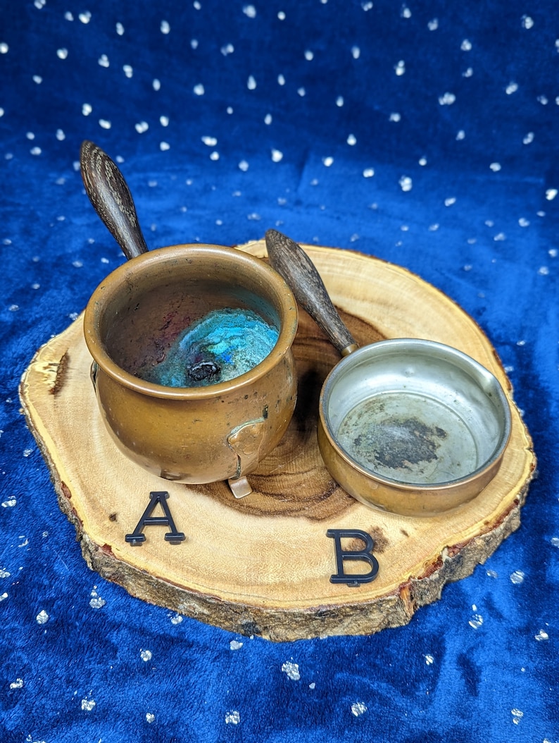 May include: Two small copper pots with wooden handles rest on a wooden slice. One pot is filled with blue and green material. Black letters A and B are placed near the pots.