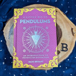 May include: A Little Bit of Pendulums book with a pink cover and gold accents, featuring a pendulum illustration. The book rests on a wooden slice against a dark blue, textured background. Text includes "An Introduction to Pendulum Divination" and the author's name.