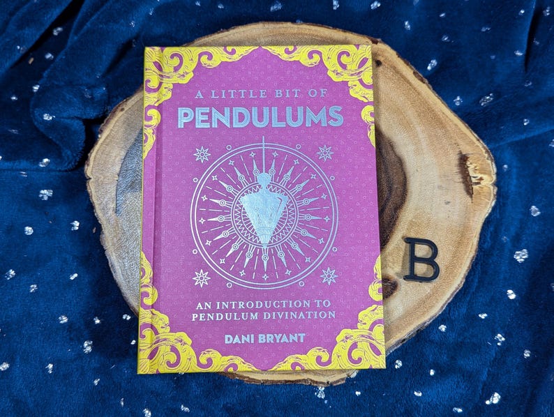 May include: A Little Bit of Pendulums book with a pink cover and gold accents, featuring a pendulum illustration. The book rests on a wooden slice against a dark blue, textured background. Text includes "An Introduction to Pendulum Divination" and the author's name.
