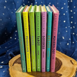 May include: A collection of colorful books titled "A Little Bit Of..." on topics like Astrology, Pendulums, Runes, Tarot, Goddess, Dreams, and Numerology. The books have different colored spines and are arranged on a wooden surface.