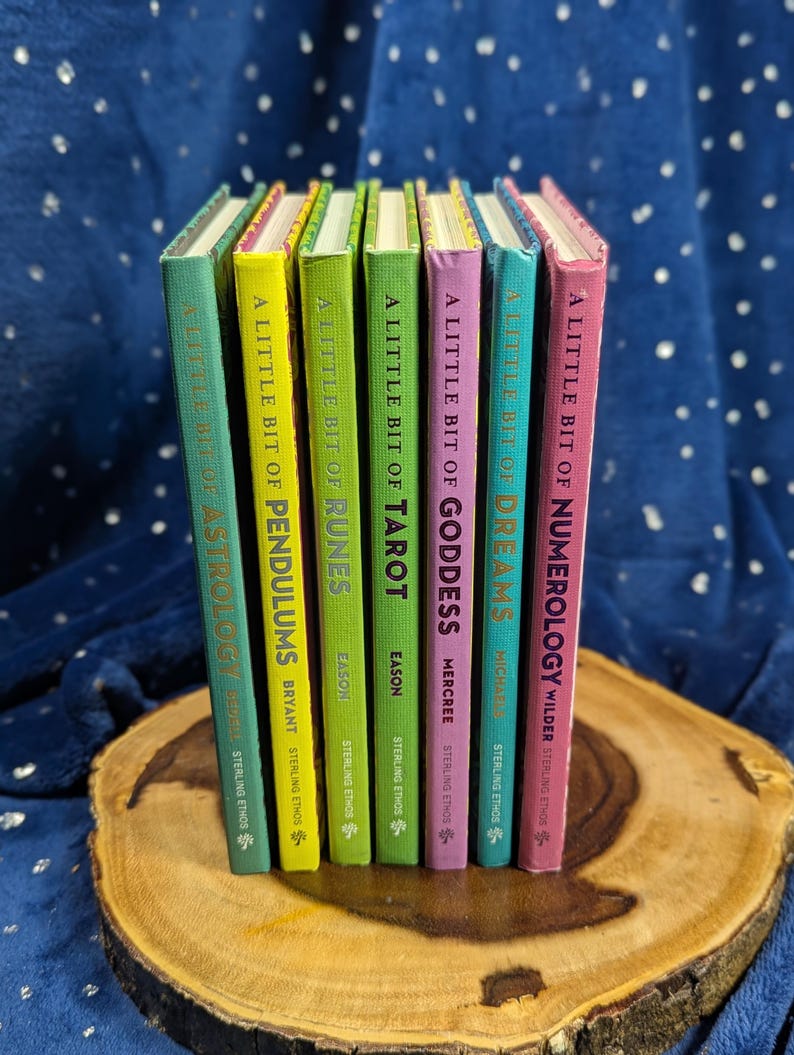 May include: A collection of colorful books titled "A Little Bit Of..." on topics like Astrology, Pendulums, Runes, Tarot, Goddess, Dreams, and Numerology. The books have different colored spines and are arranged on a wooden surface.