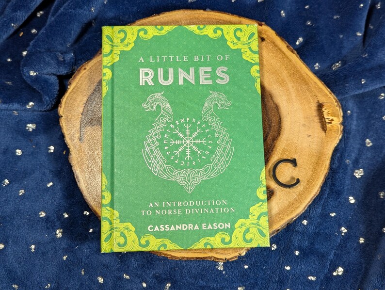 May include: A green book titled "A Little Bit of Runes" with white lettering and illustrations, including a Viking symbol. The book is on a wooden slice with a dark blue background.