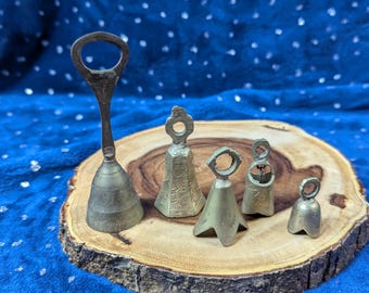 May include: Five brass bells of varying sizes, with one bell having a bottle opener on top, arranged on a wooden slice.