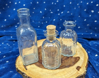May include: Three clear glass bottles with cork stoppers, two of the bottles have a textured surface, the bottles are sitting on a wooden slice.