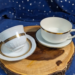 May include: Two white teacups with gold trim and matching saucers on a wooden surface. The teacups are empty and ready for use.