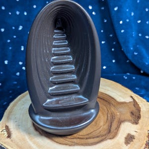 May include: A black ceramic incense burner shaped like a cave with a set of stairs leading to a small opening at the top. The burner is sitting on a wooden base.