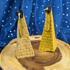 May include: Two pyramid-shaped candles with brick-like textures. One is tan with an eye symbol, the other is yellow with a triangle symbol. Each has a black heart tag with the text "nugmAgicks". They sit on a wooden slice against a blue background.
