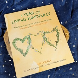 May include: A book titled "A Year of Living Kindly" with text and floral heart illustrations. The cover is a light tan color, with green and white floral accents. The book is on a wooden surface, set against a dark blue background.