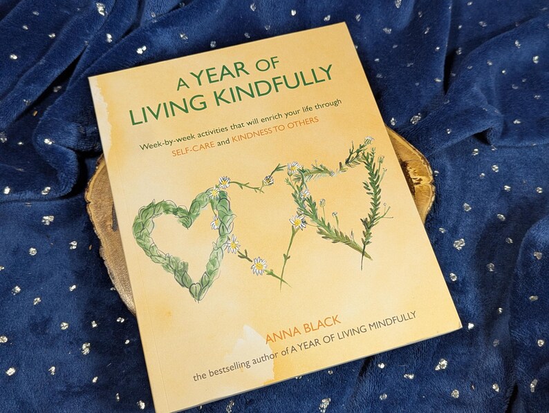May include: A book titled "A Year of Living Kindly" with text and floral heart illustrations. The cover is a light tan color, with green and white floral accents. The book is on a wooden surface, set against a dark blue background.
