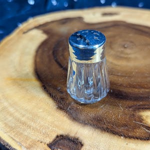 May include: A small, clear glass salt or pepper shaker featuring a vertical ribbed pattern on the body and a polished, reflective silver-toned metal cap with a series of small holes on the top for dispensing seasonings.