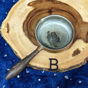 May include: A small copper saucepan with a wooden handle rests on a wooden slice. The interior of the pan shows signs of use. A black letter "B" is placed near the pan on a blue surface.
