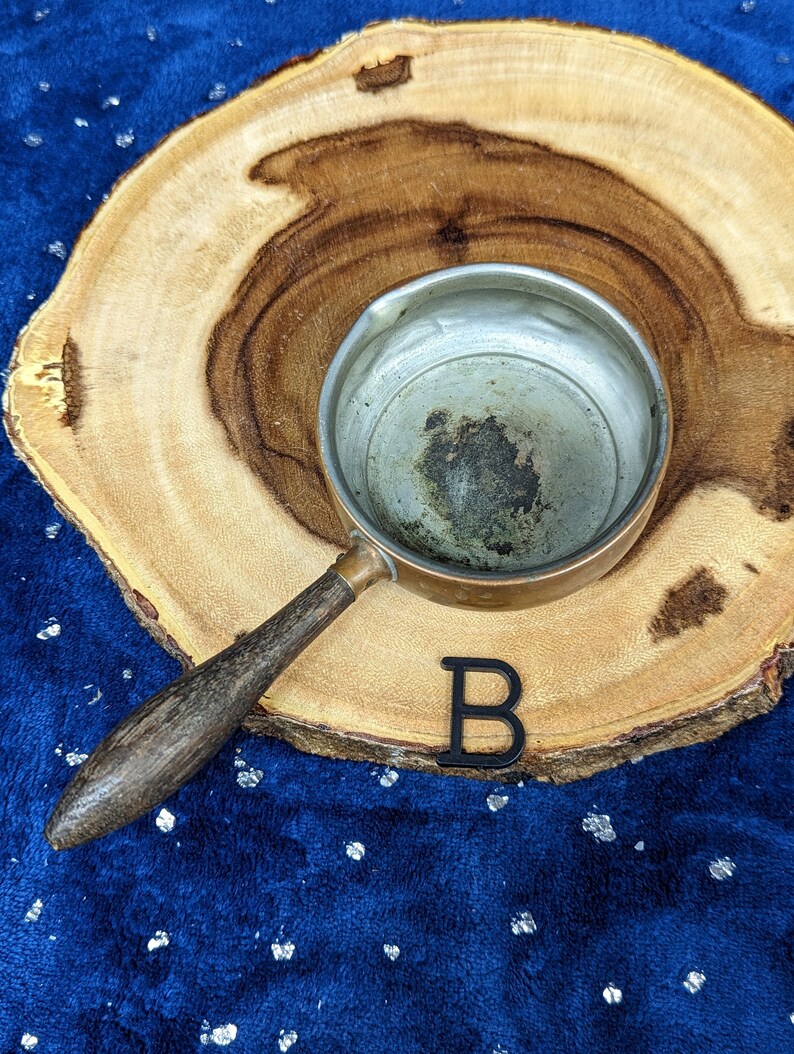May include: A small copper saucepan with a wooden handle rests on a wooden slice. The interior of the pan shows signs of use. A black letter "B" is placed near the pan on a blue surface.