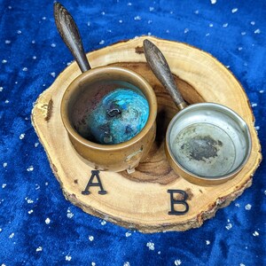 May include: Two small copper pots with wooden handles rest on a wooden slice. One pot contains a blue and green substance, while the other is empty. The pots are labeled A and B with black letters.