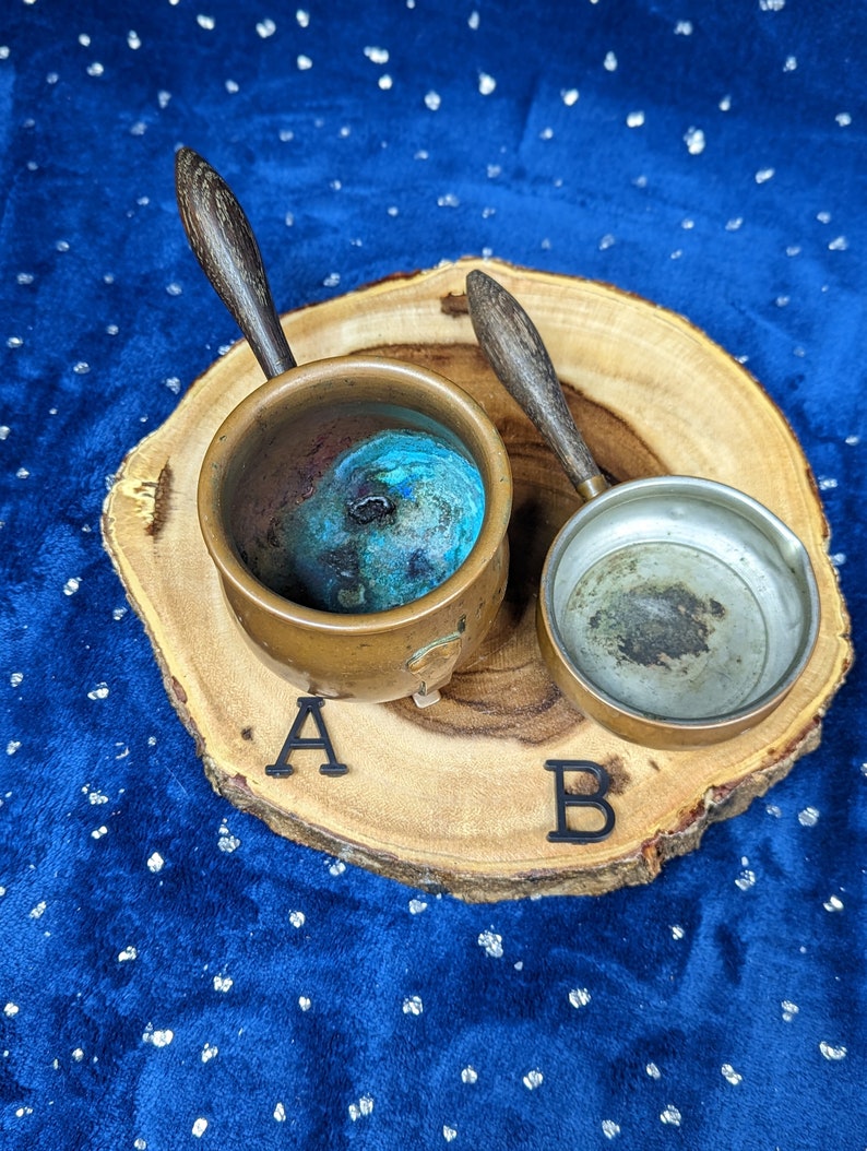 May include: Two small copper pots with wooden handles rest on a wooden slice. One pot contains a blue and green substance, while the other is empty. The pots are labeled A and B with black letters.