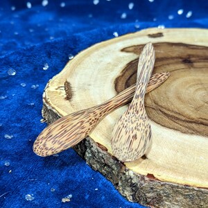 May include: Two wooden spoons with a natural wood grain pattern are displayed on a wooden slice. The spoons are brown and have a slightly curved shape.