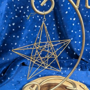 May include: A gold wire star ornament hanging from a gold metal stand. The star is a geometric design with many points and lines.