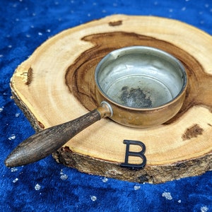 May include: A small, vintage copper saucepan with a wooden handle rests on a wooden slice. The interior of the pan is silver. A black letter "B" is placed near the pan. The background is a dark blue fabric.
