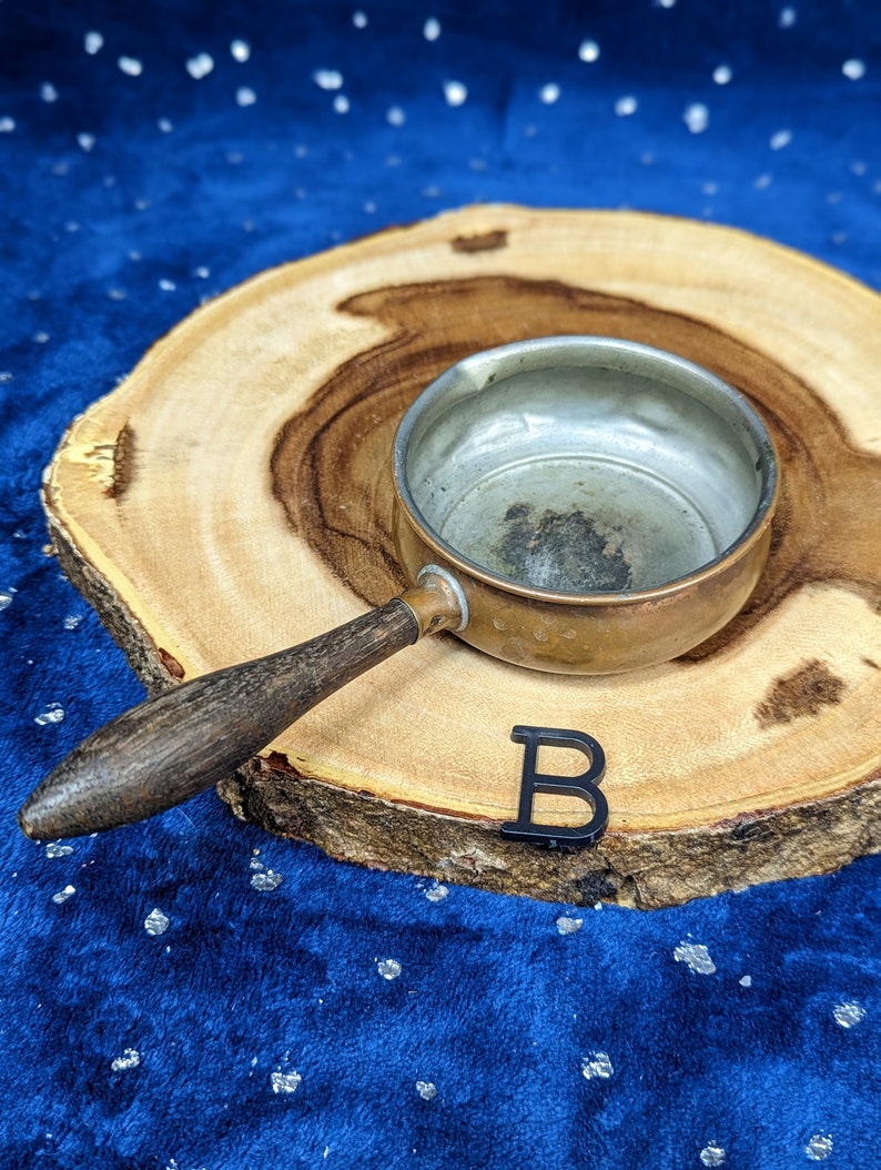 May include: A small, vintage copper saucepan with a wooden handle rests on a wooden slice. The interior of the pan is silver. A black letter "B" is placed near the pan. The background is a dark blue fabric.