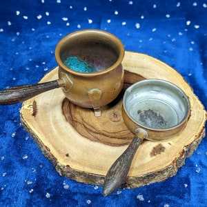 May include: Two small copper pots with wooden handles rest on a wooden slice against a blue background. One pot is open, revealing a turquoise interior. The other pot is smaller and has a silver interior.