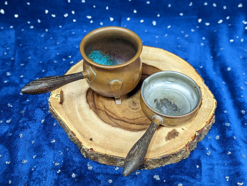 May include: Two small copper pots with wooden handles rest on a wooden slice against a blue background. One pot is open, revealing a turquoise interior. The other pot is smaller and has a silver interior.