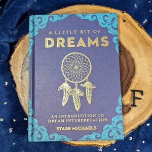 May include: A Little Bit of Dreams book with a dreamcatcher design on the cover. The book is purple with gold lettering and teal accents. The book is on a wooden slice.