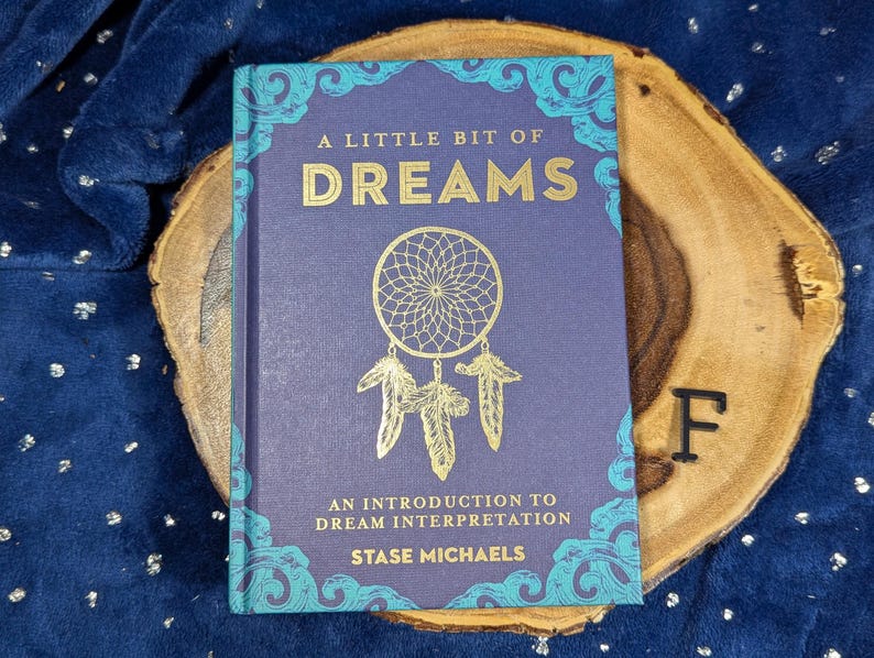 May include: A Little Bit of Dreams book with a dreamcatcher design on the cover. The book is purple with gold lettering and teal accents. The book is on a wooden slice.