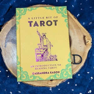 May include: A yellow book titled "A Little Bit of Tarot" with green and purple accents. The cover features an illustration of a figure with raised arms. The book is an introduction to reading tarot by Cassandra Eason.