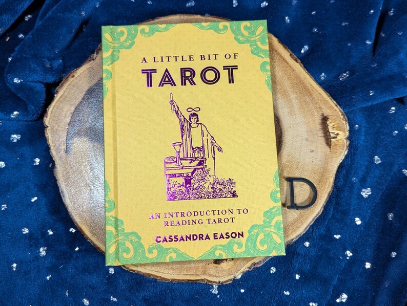 May include: A yellow book titled "A Little Bit of Tarot" with green and purple accents. The cover features an illustration of a figure with raised arms. The book is an introduction to reading tarot by Cassandra Eason.