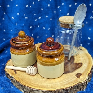 May include: Two brown and white ceramic jars with lids, a wooden honey dipper, and a clear glass jar with a cork lid and a silver spoon on a wooden slice.