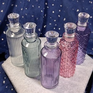 May include: Five glass bottles with clear faceted stoppers. Three bottles feature vertical ribbed textures in clear, sage green, and light purple. Two bottles feature diamond-patterned textures in pink and deep purple, arranged on a light gray base.
