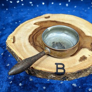 May include: A small, vintage copper saucepan with a wooden handle rests on a wooden slice. The interior of the pan shows signs of use. A black letter "B" is placed near the pan. The background is a dark blue fabric with white specks.
