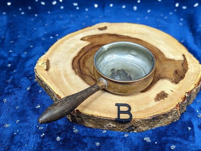 May include: A small, vintage copper saucepan with a wooden handle rests on a wooden slice. The interior of the pan shows signs of use. A black letter "B" is placed near the pan. The background is a dark blue fabric with white specks.