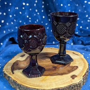 May include: Two dark red glass goblets with a textured design, sitting on a wooden slice.