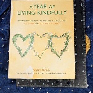 May include: A book titled "A Year of Living Kindly" with floral heart illustrations. The cover is light tan with green and white floral accents. The book is on a blue surface with a ruler next to it.