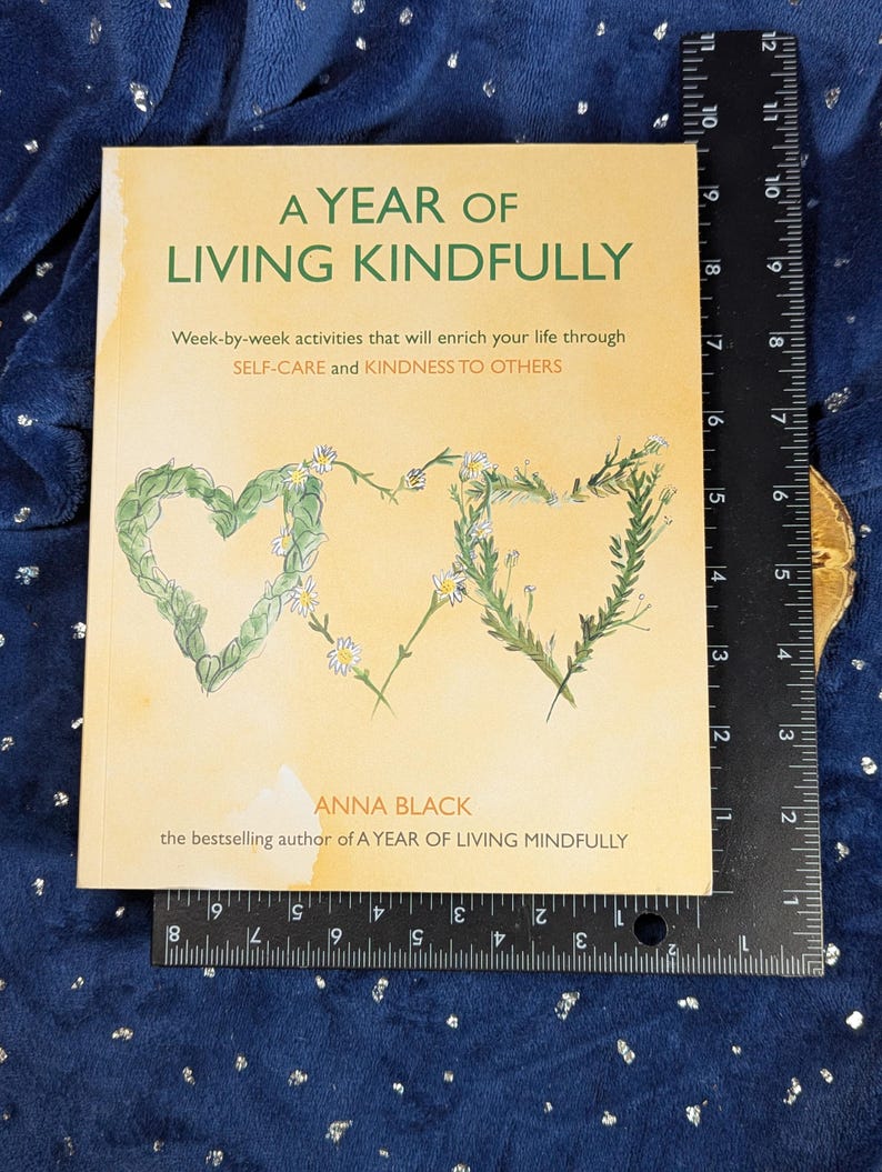 May include: A book titled "A Year of Living Kindly" with floral heart illustrations. The cover is light tan with green and white floral accents. The book is on a blue surface with a ruler next to it.