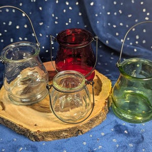 May include: Four glass candle holders with metal handles. The holders are clear, red, and green. They are sitting on a wooden slice.