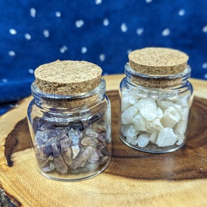 May include: Two small glass jars with cork lids, filled with small, colorful crystals. The jar on the left contains purple crystals, and the jar on the right contains white crystals.