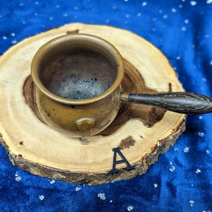 May include: A small, antique copper pot with a wooden handle. The pot is round with a slightly flared rim and a small spout. The handle is dark brown and attached with metal rivets. The pot sits on a round wooden slice against a blue background.