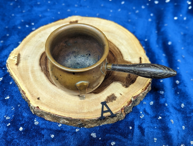 May include: A small, antique copper pot with a wooden handle. The pot is round with a slightly flared rim and a small spout. The handle is dark brown and attached with metal rivets. The pot sits on a round wooden slice against a blue background.