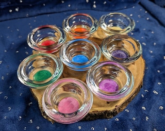 May include: A wooden tray with eight small glass cups, each containing a different colored sand. The colors are red, orange, yellow, green, blue, purple, pink, and white.